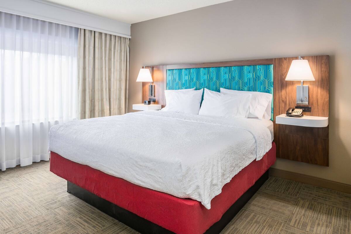 A hotel room with a large bed, two bedside tables with lamps, a wooden headboard, and a view of the outside through sheer curtains.