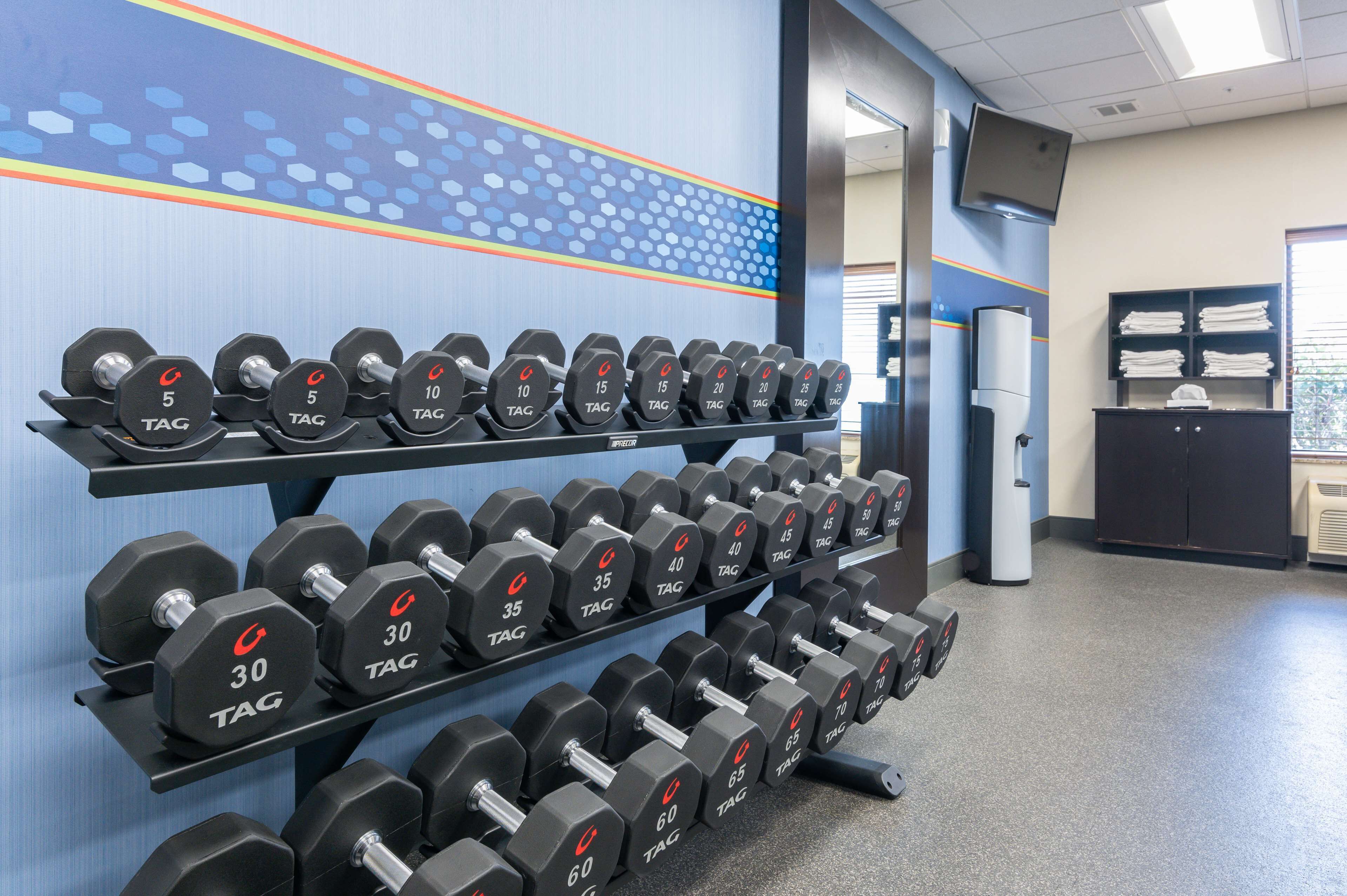 A gym with multiple rows of dumbbells on racks, a television mounted on the wall, and a water cooler.