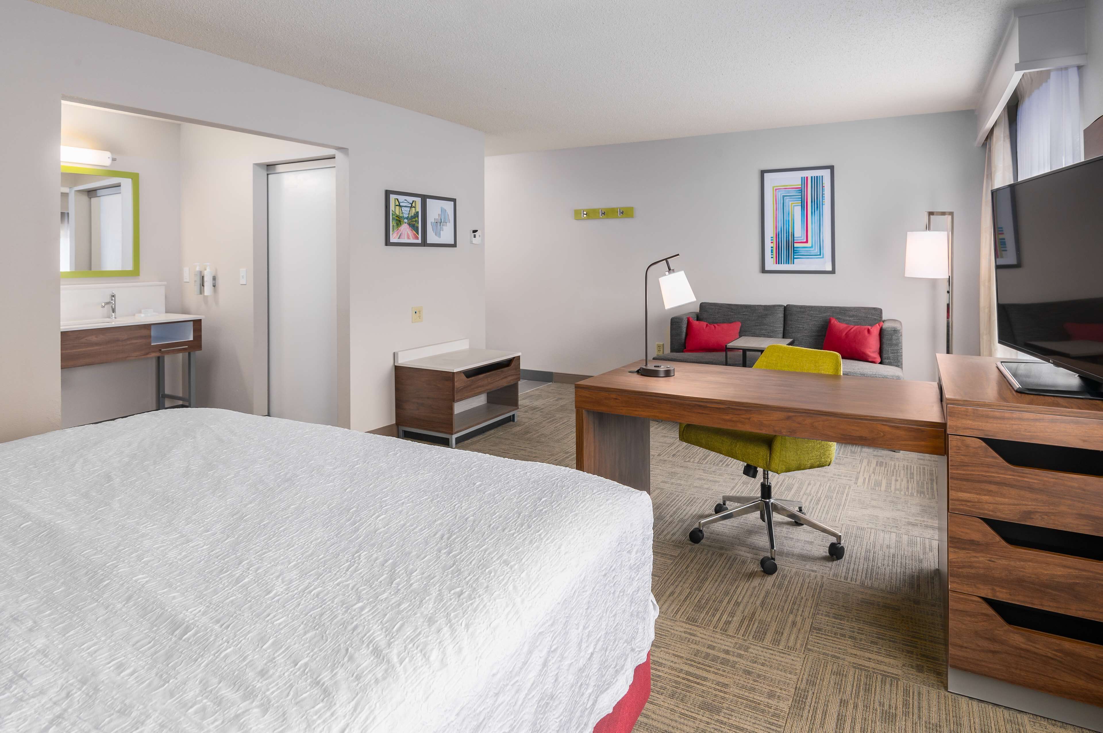 A hotel room with a large bed, a desk with a chair, a sofa with red cushions, a TV, and a bathroom with a mirror and sink.