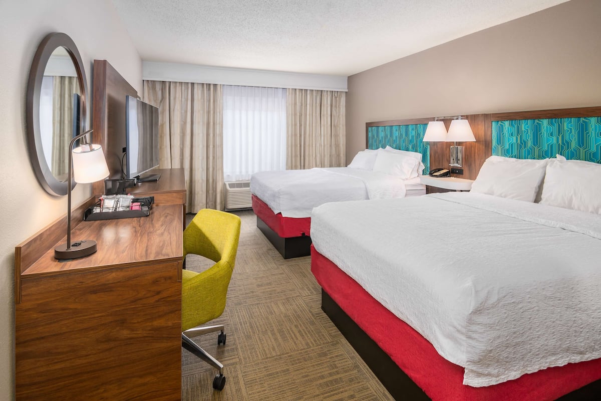 A hotel room with two beds, a desk with a lamp, a chair, and a large mirror.