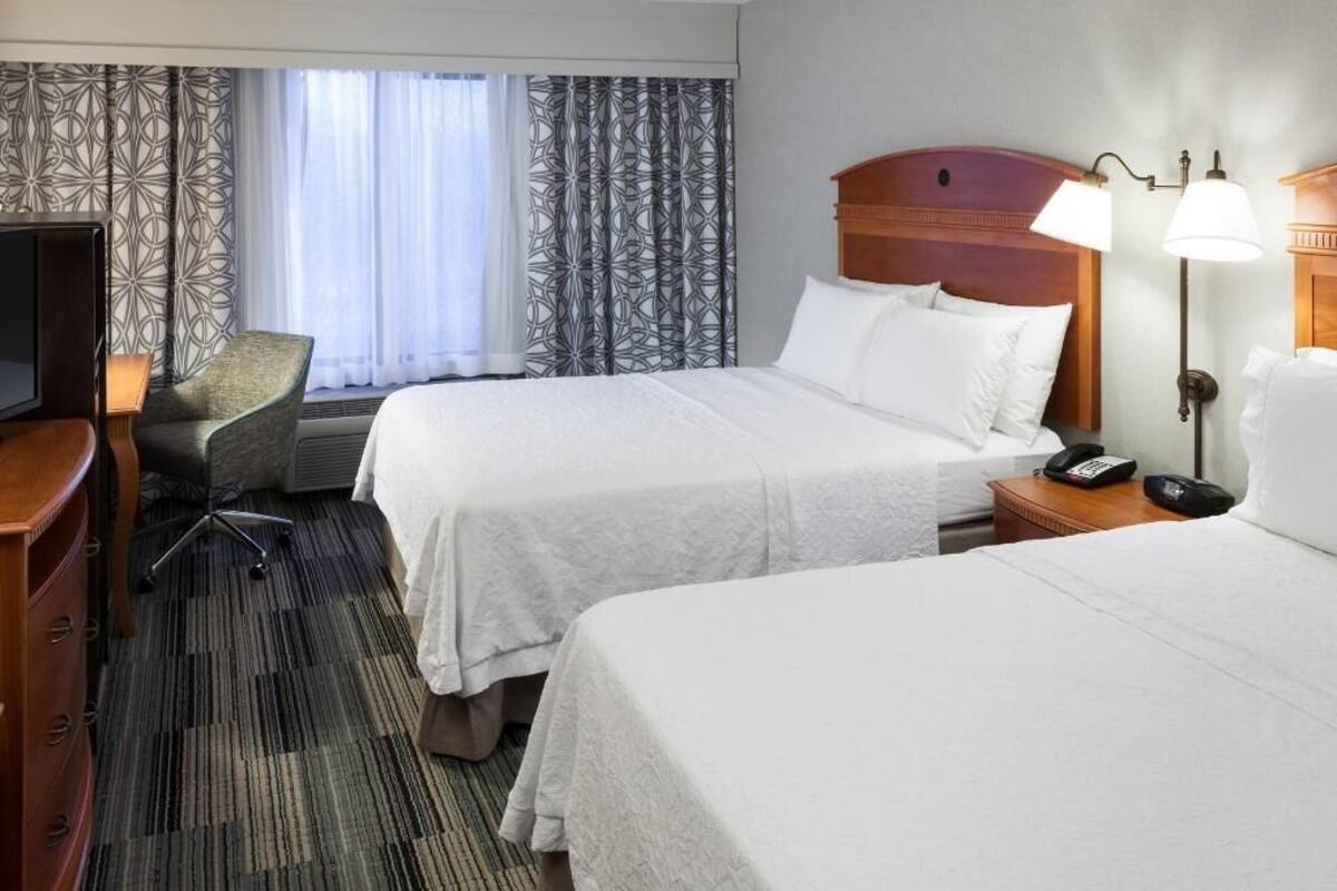 A hotel room with two beds, a desk, a chair, a TV, and a lamp.