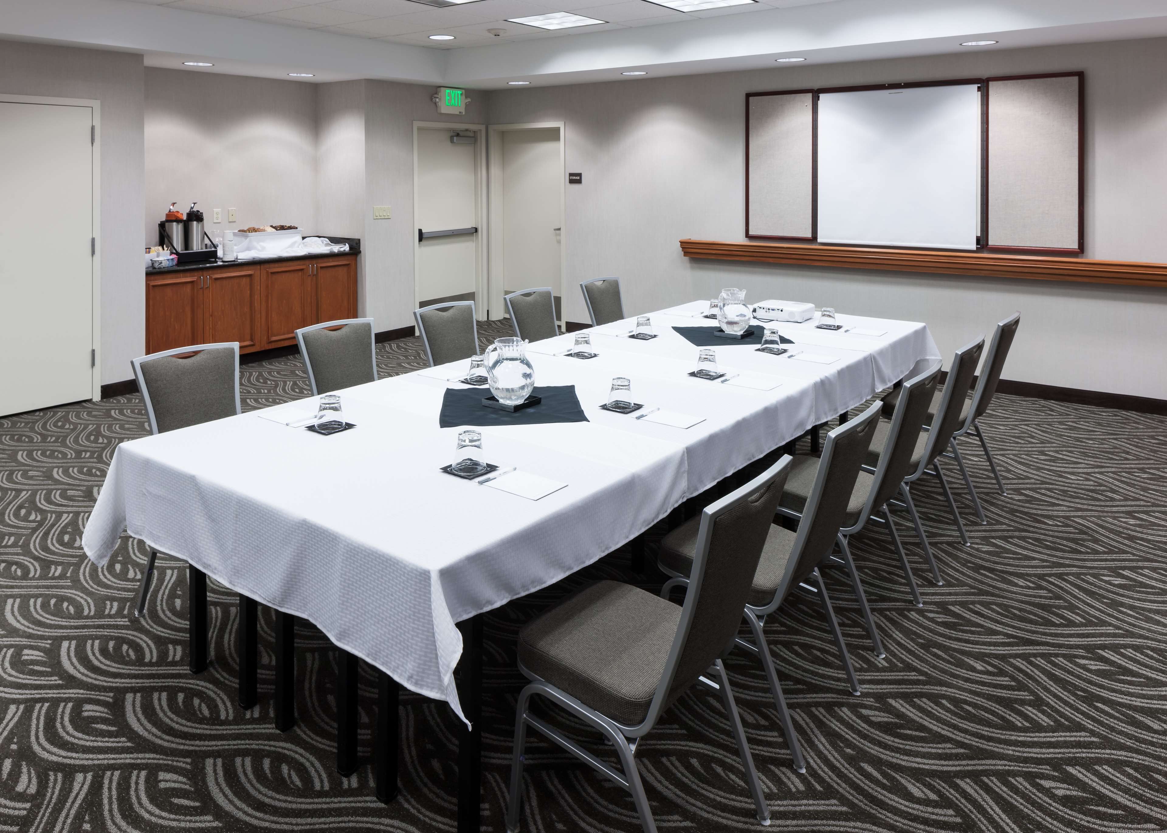 Un salón de conferencias con una larga mesa, sillas, una pantalla de proyector y una zona de buffet.