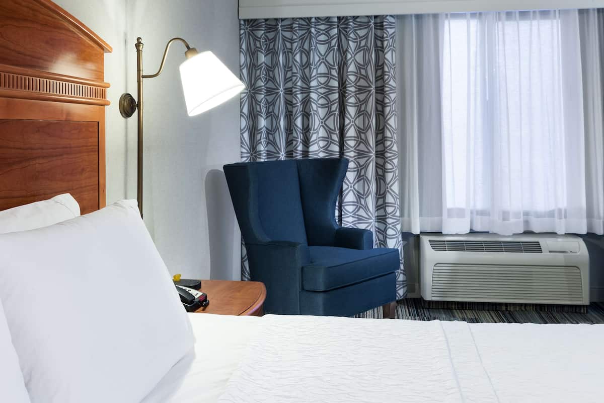 A hotel room with a bed, a blue armchair, a desk with a lamp, patterned curtains, and an air conditioning unit.