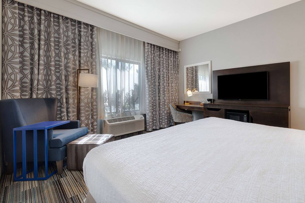 A hotel room with a large bed, a desk with a flat-screen TV, a chair, and a desk lamp.