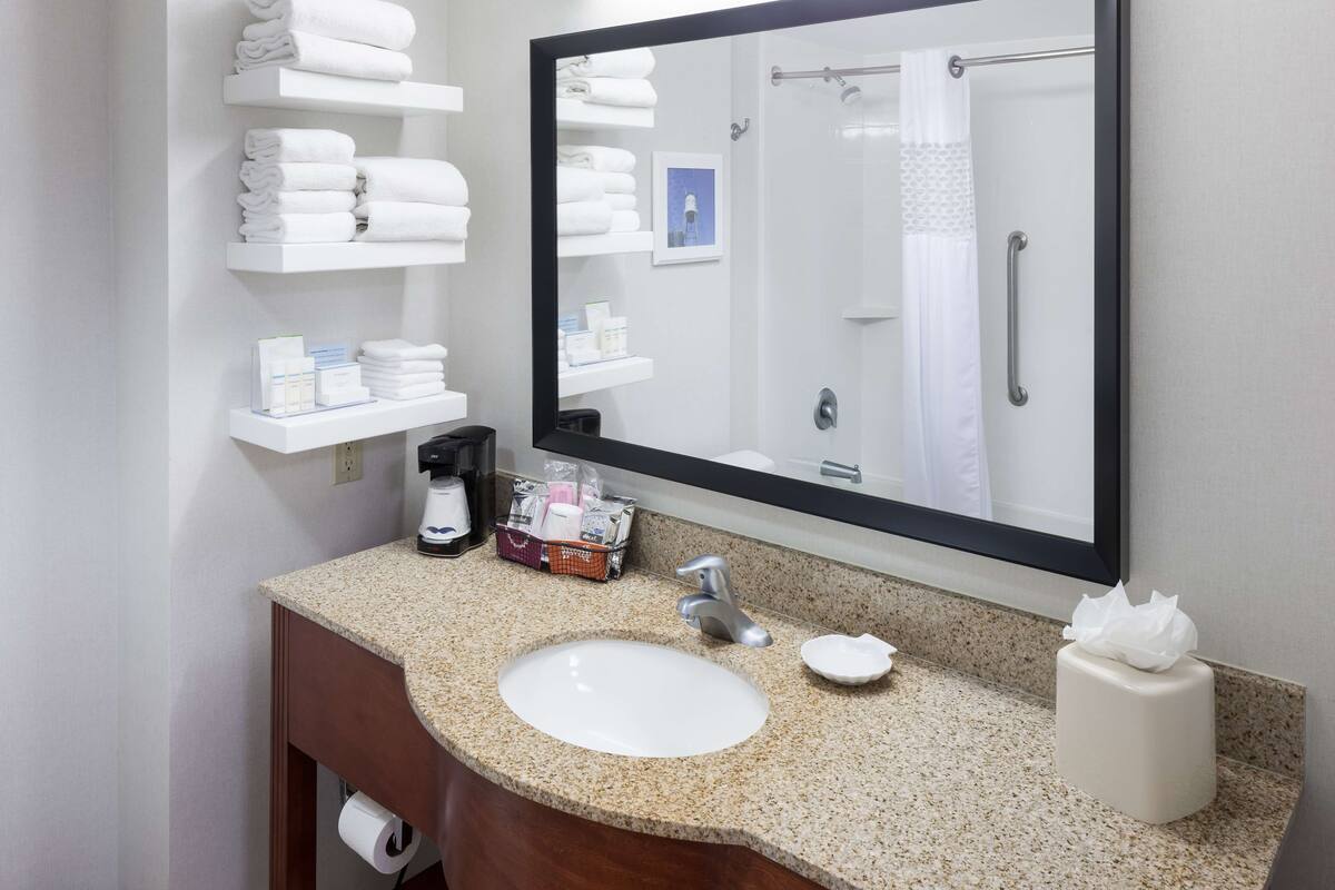 A bathroom with a granite countertop, a white sink, a coffee maker, and a mirror reflecting a shower area.