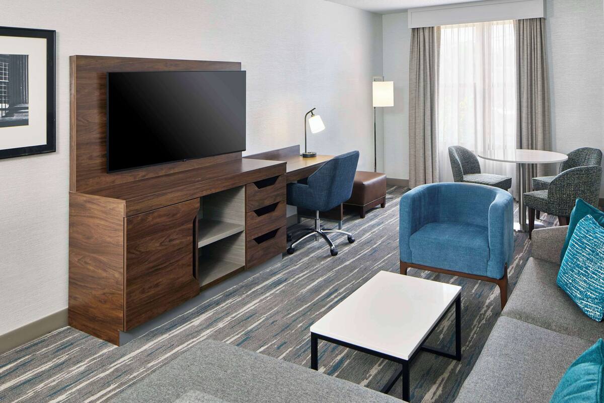 A hotel room with a flat-screen TV, a wooden desk with drawers, a blue armchair, a small table, and a round coffee table.