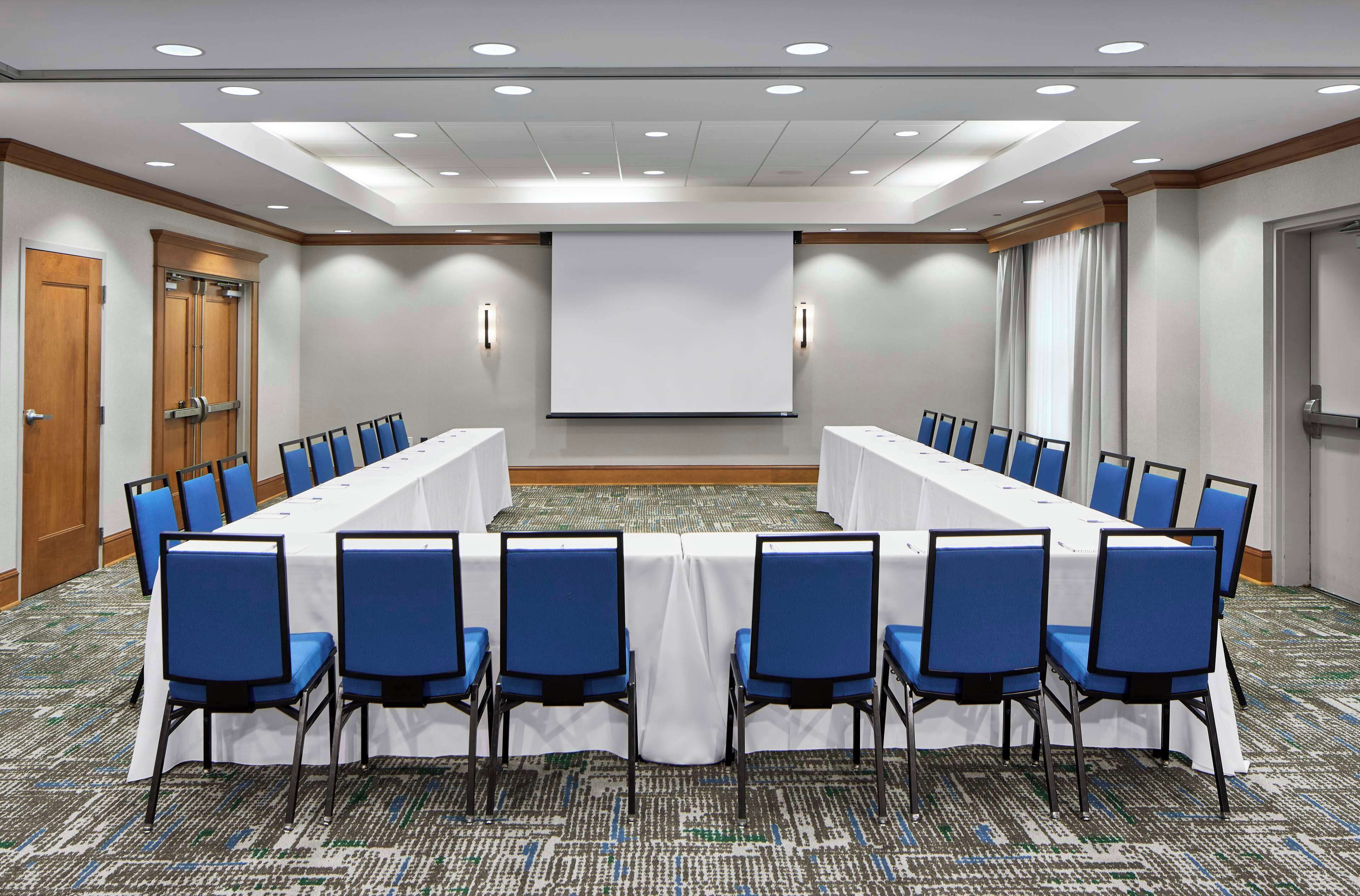 Une salle de conférence avec une longue table, des chaises bleues, un écran de projection et un sol recouvert de moquette.