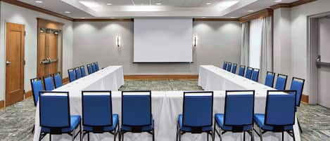 A conference room with a long table, blue chairs, a projector screen, and a carpeted floor.