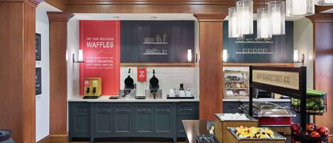 A hotel breakfast area with a red sign advertising waffles, a coffee station, and a fruit display.