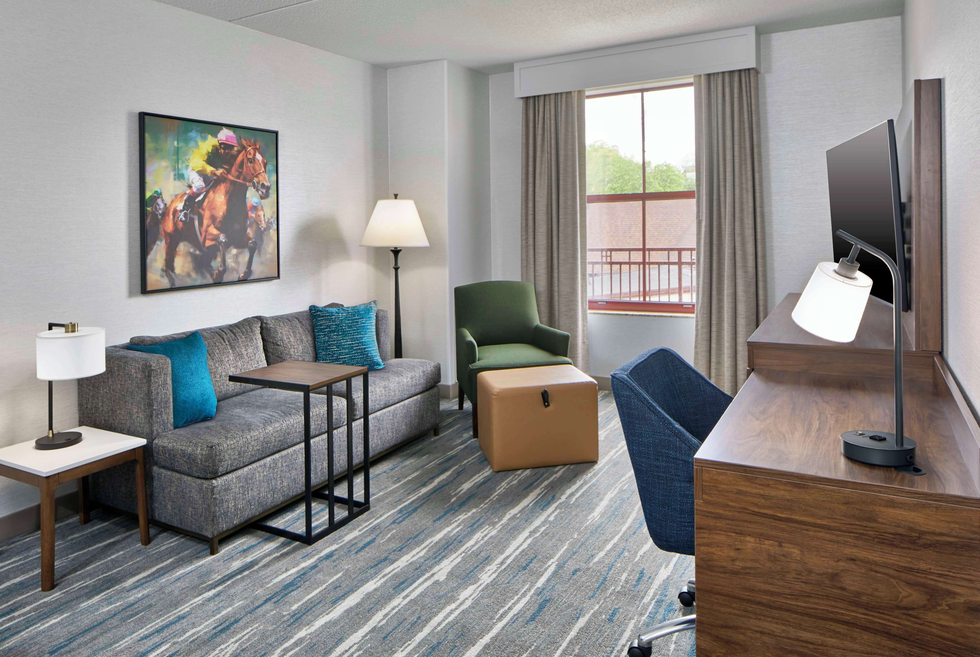 A hotel room with a gray sofa, a green armchair, a wooden desk, a flat-screen TV, a lamp, and a painting of horses on the wall.
