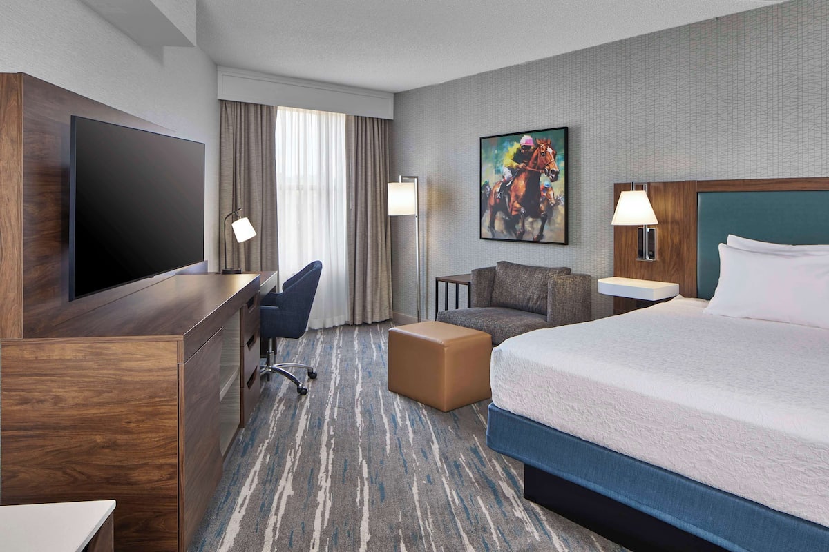 A hotel room with a large bed, a desk with a flat-screen TV, a sofa, a small ottoman, and a painting on the wall.