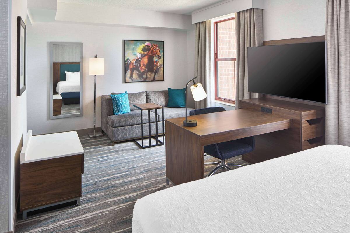 A hotel room with a bed, a desk with a lamp, a sofa with cushions, a TV, and a painting on the wall.