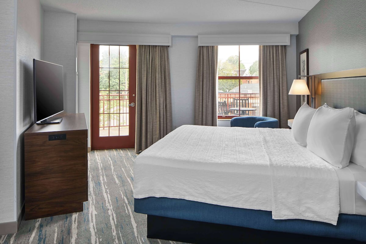A hotel room with a large bed, a television on a wooden cabinet, a desk, and a view of the outside through a glass door.