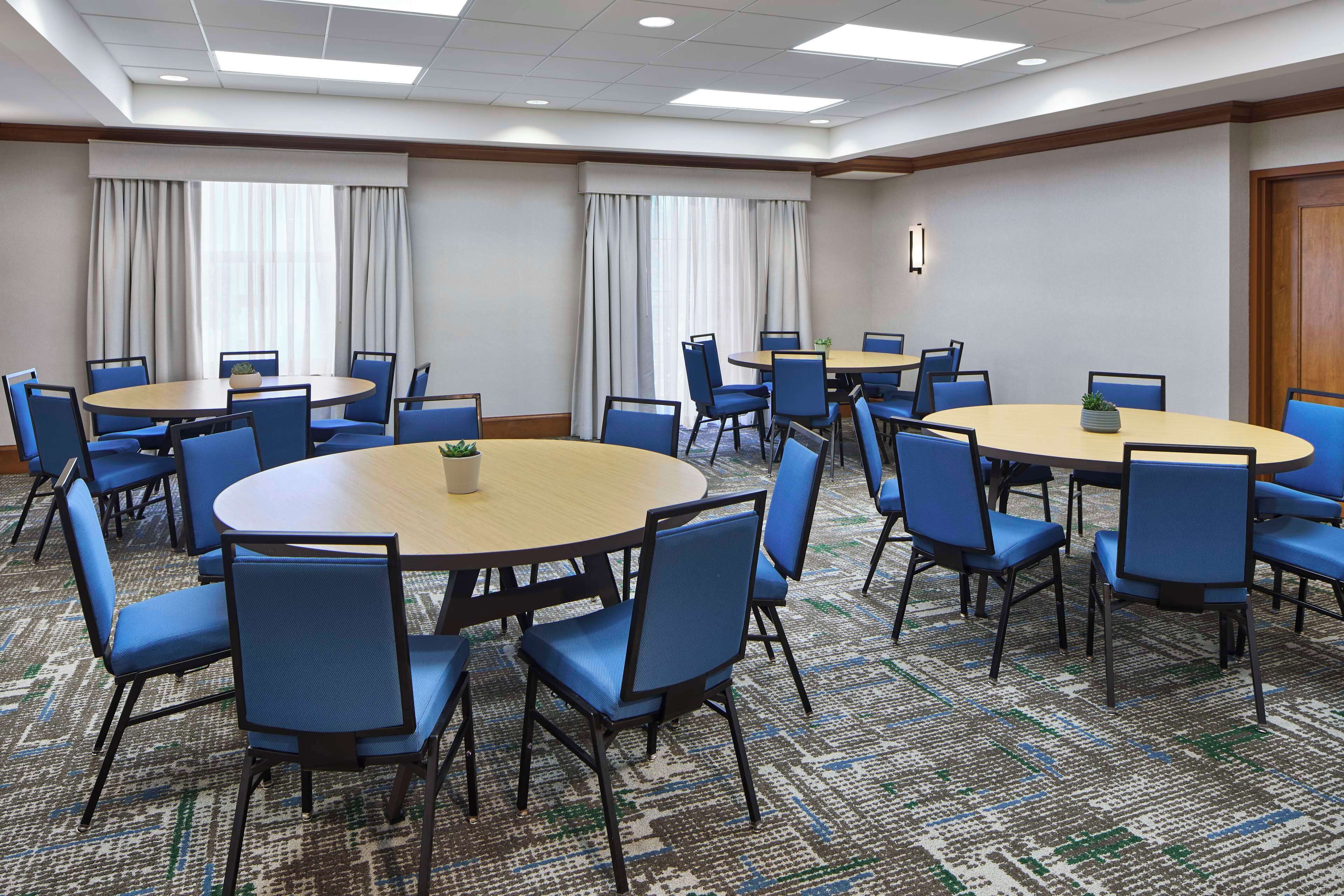 A conference room with multiple round tables and blue chairs, carpeted floor, and large windows with curtains.