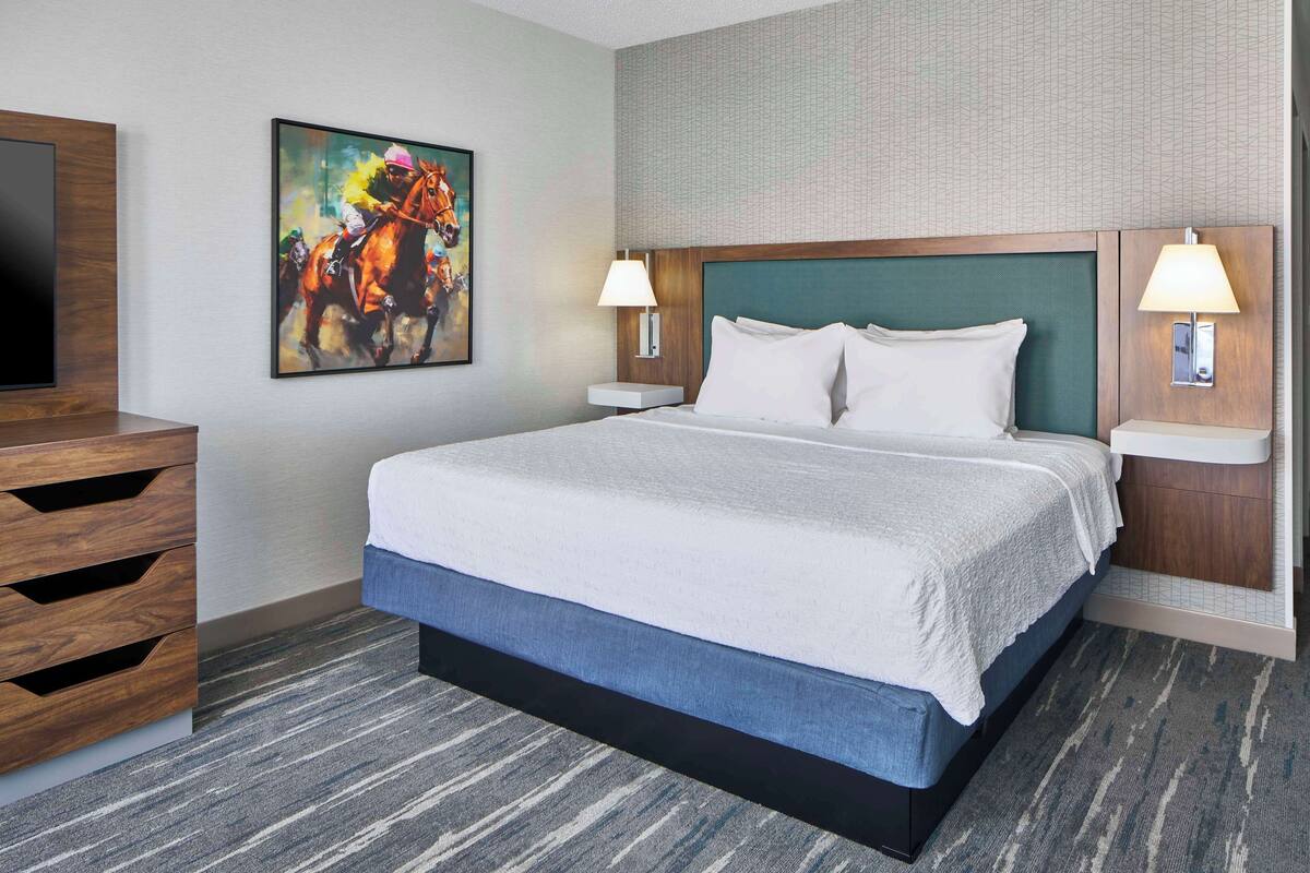A hotel room with a large bed, two bedside lamps, a mounted TV, and a painting of a horse race.