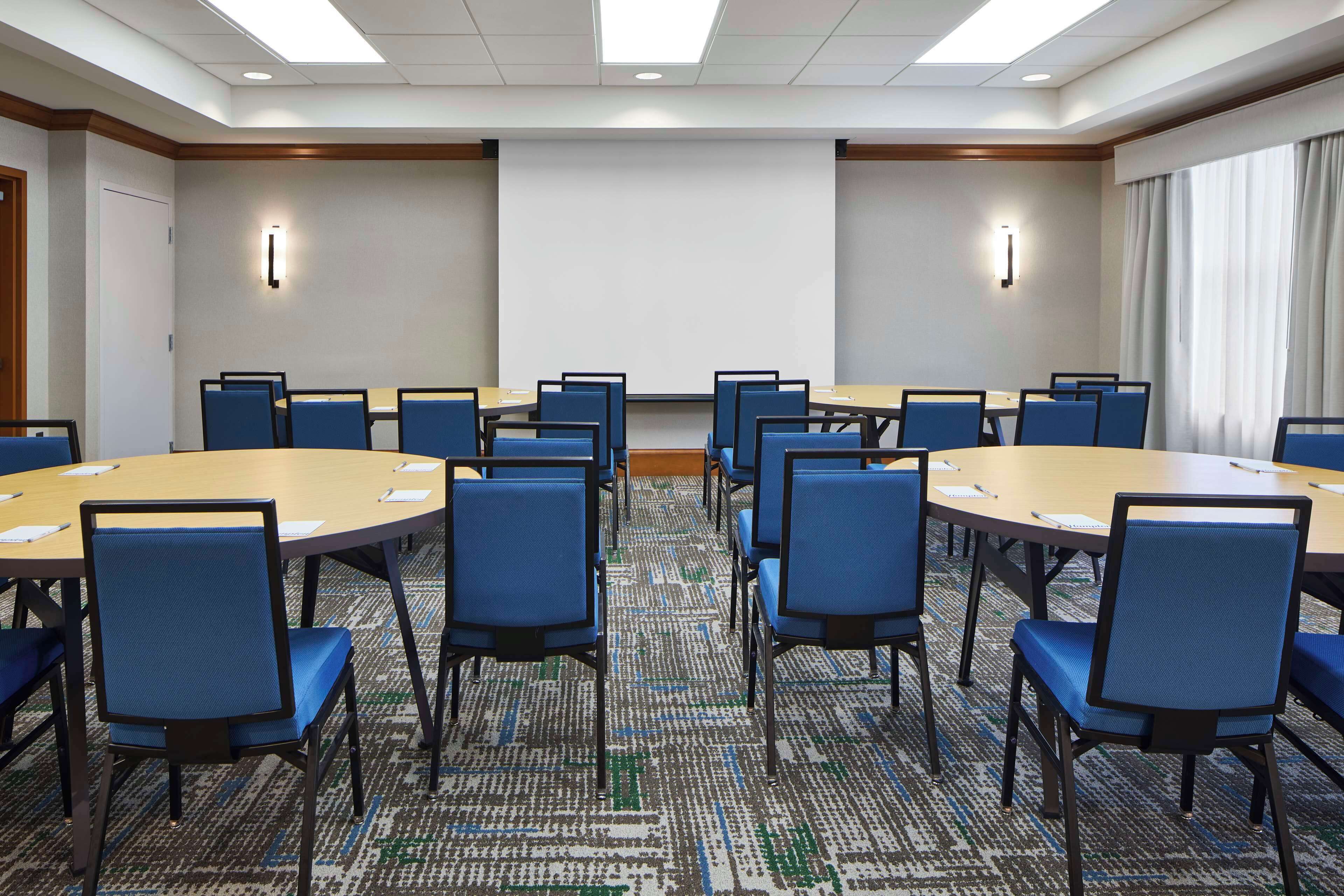 A conference room with multiple tables and chairs, a projector screen, and a carpeted floor.