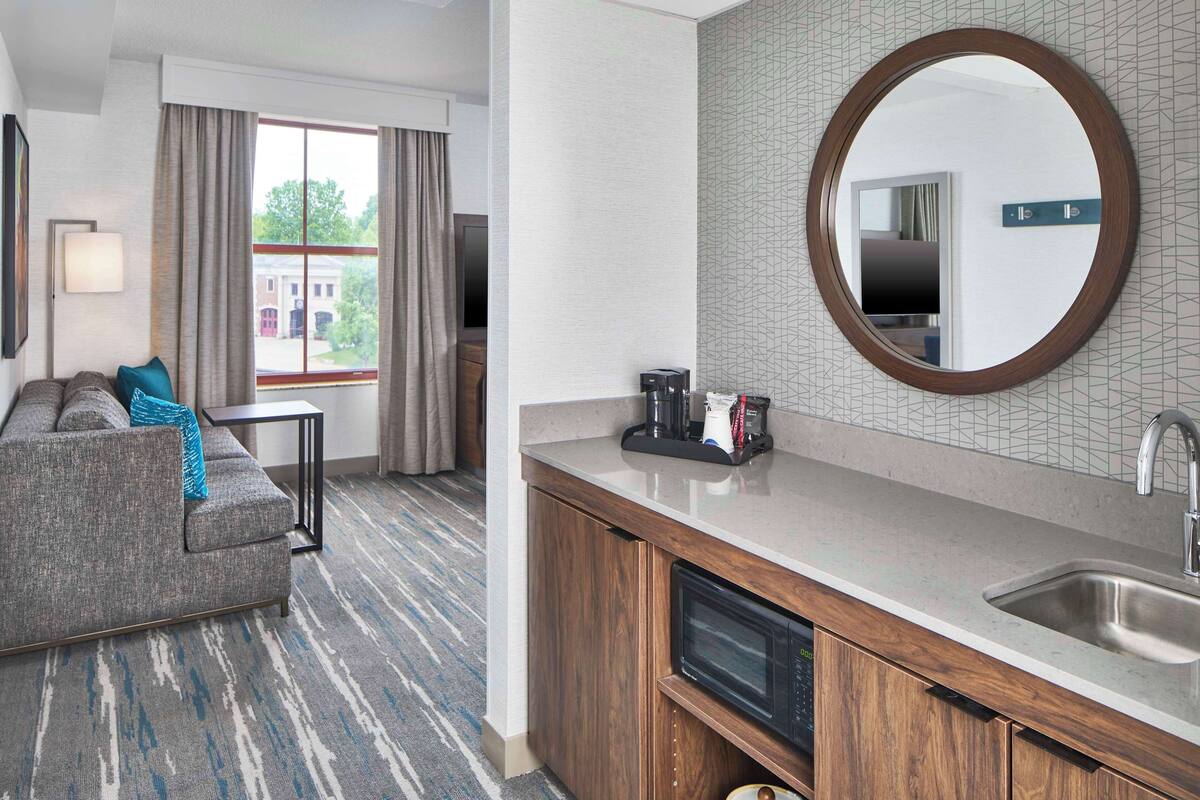 A hotel room with a sofa, a small table, a microwave, a sink, and a large mirror.