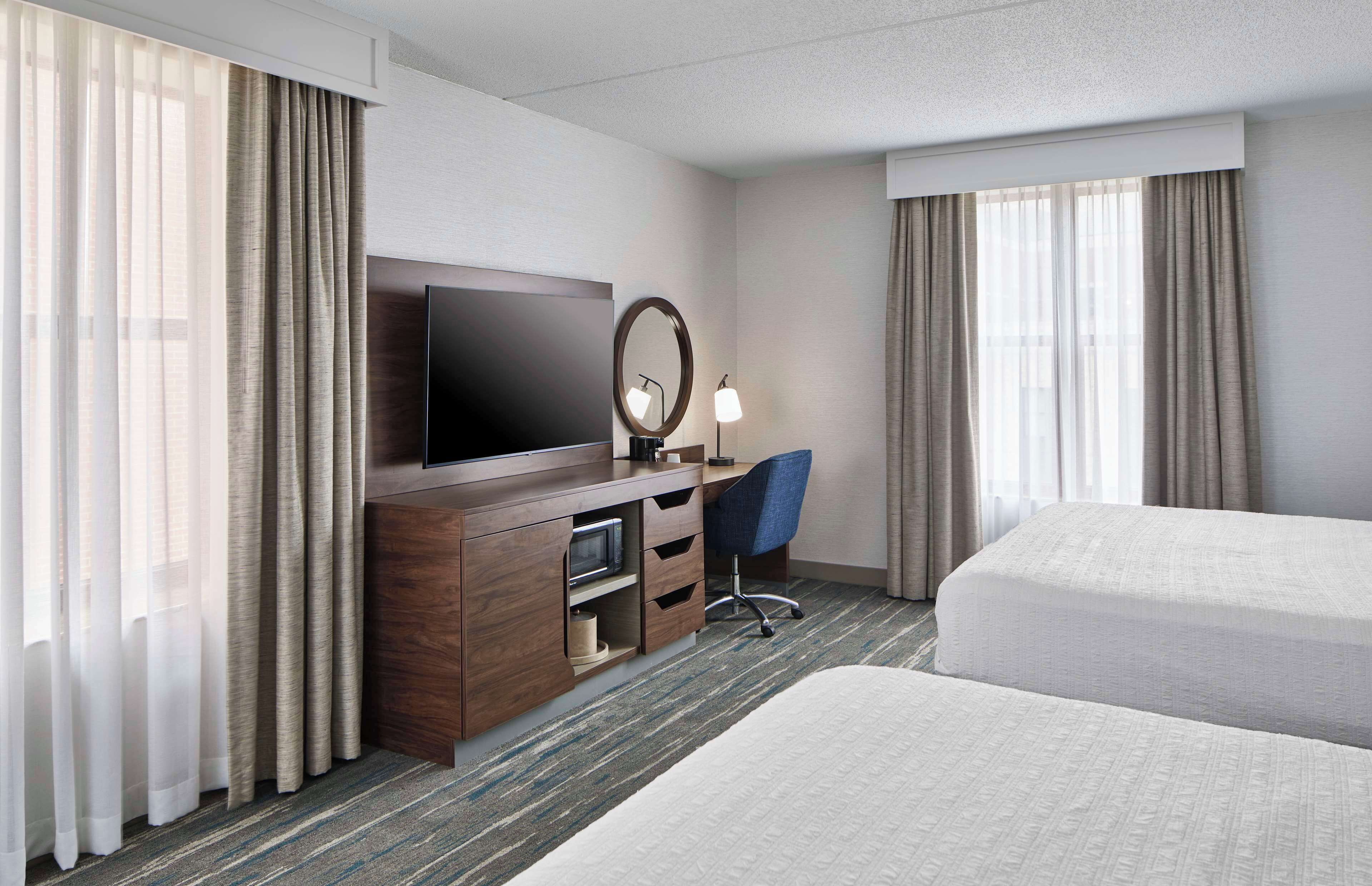 Une chambre d’hôtel avec un lit, un bureau avec une télévision, une chaise et une grande fenêtre avec des rideaux.