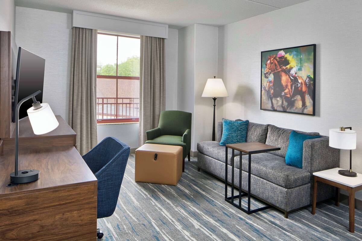 A hotel room with a flat-screen TV, a green armchair, a grey sofa with blue cushions, a small table, a lamp, and a painting of horses.