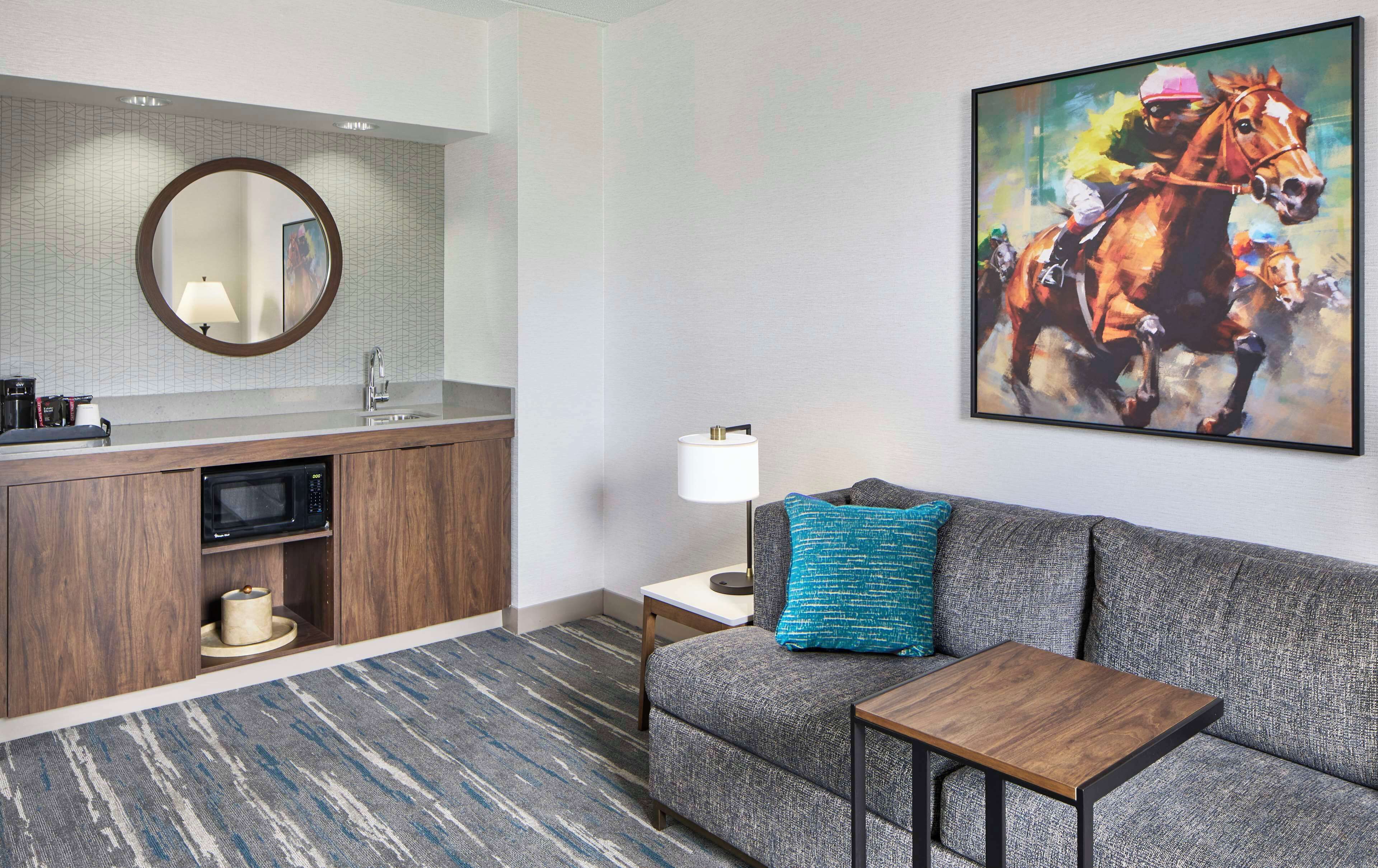 A modern hotel room with a kitchenette, a sofa with a blue pillow, a small table, and a large painting of a horse.