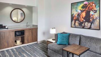 A modern hotel room with a kitchenette, a sofa with a blue pillow, a small table, and a large painting of a horse.