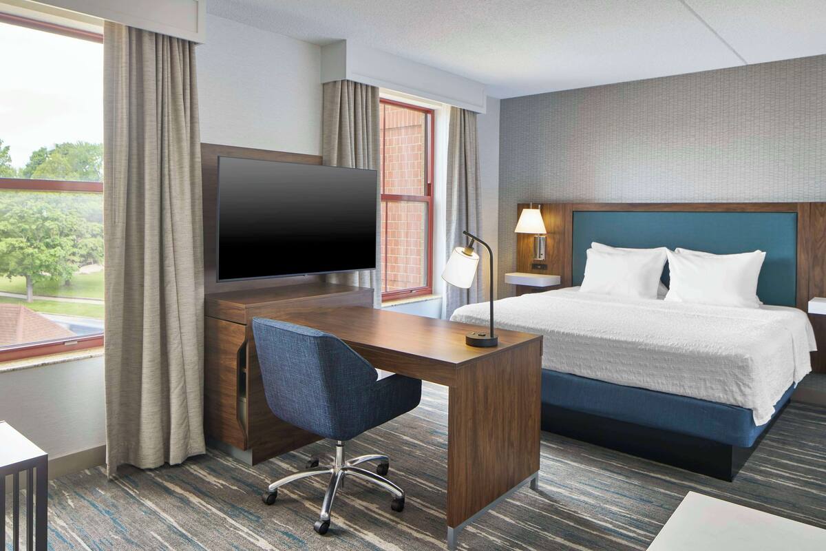 A hotel room with a bed, a desk with a flat-screen TV, a chair, and a window with curtains.