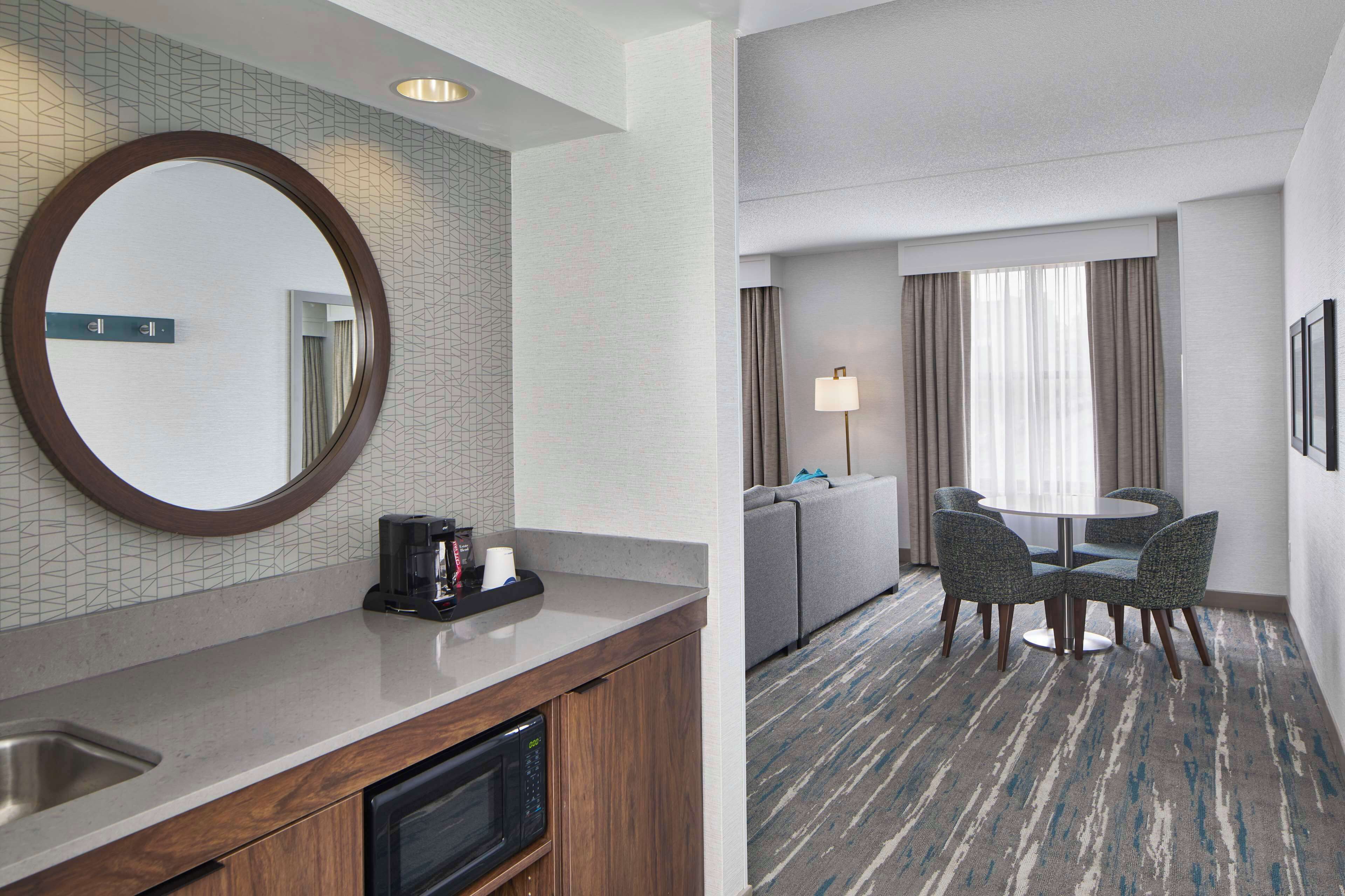 Una habitación de hotel con cocina, una mesa de comedor con sillas, un espejo grande y una lámpara.