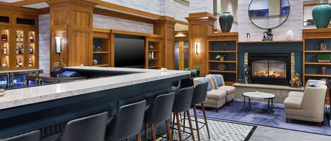 A modern bar area with a marble countertop, bar stools, and a television. There are shelves with bottles and decorative items, and a fireplace with seating around it.