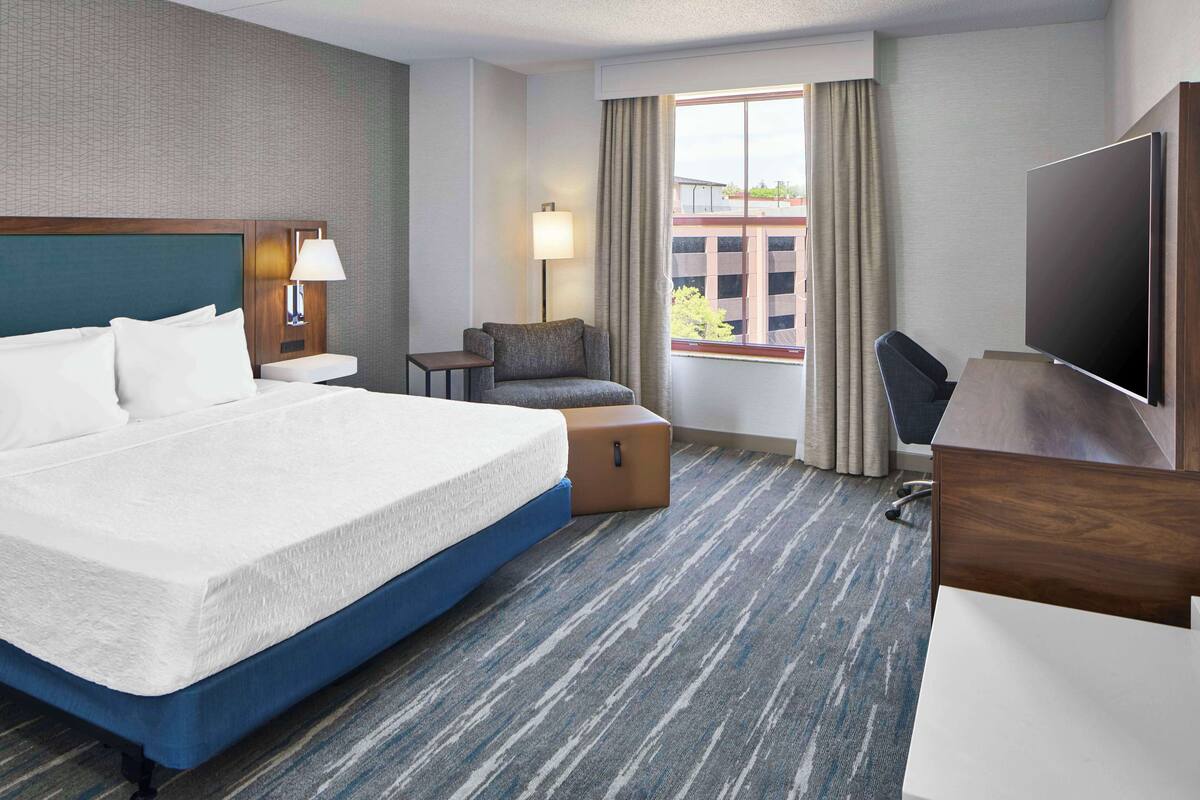 A hotel room with a large bed, a desk, a chair, a TV, a lamp, and a window with curtains.