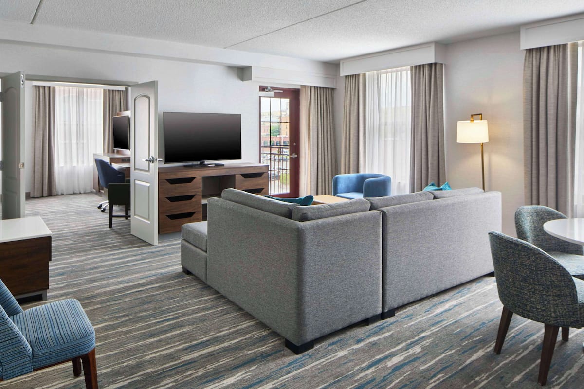 A hotel room with a large gray sofa, a television, a desk with a chair, and a dining area with a table and chairs.