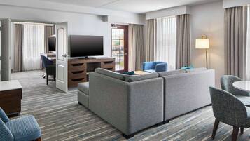 Una habitación de hotel con un sofá grande de color gris, un televisor, un escritorio con una silla y un área de comedor con mesa y sillas.