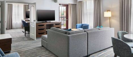 Una habitación de hotel con un sofá grande de color gris, un televisor, un escritorio con una silla y un área de comedor con mesa y sillas.