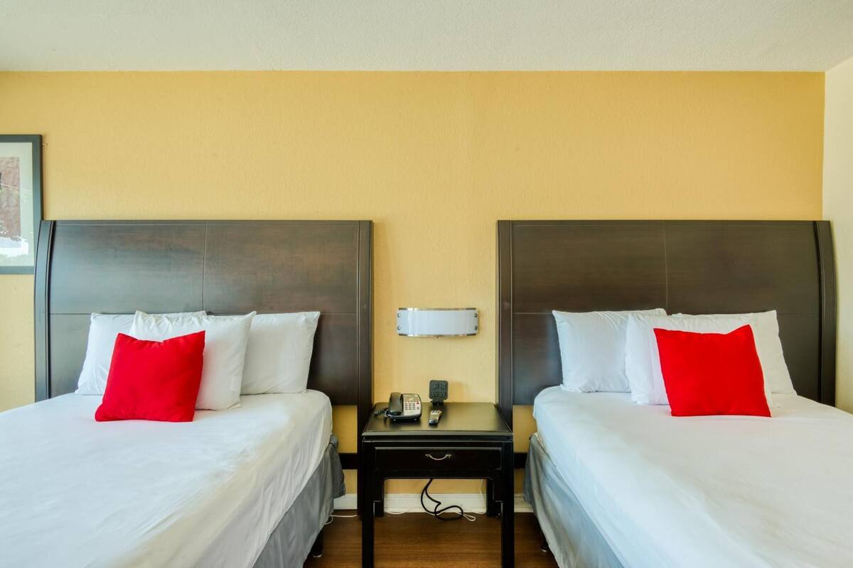 Ein Hotelzimmer mit zwei Betten, jeweils mit weißer Bettwäsche und einem roten Kopfkissen. Zwischen den Betten befindet sich ein kleiner Nachttisch mit Telefon und Lampe.