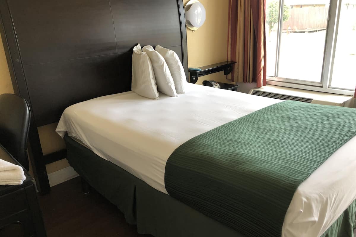 Ein Hotelzimmer mit einem großen Bett, einem dunklen Kopfteil, einem Nachttisch mit Lampe und Blick ins Freie durch ein Fenster.
