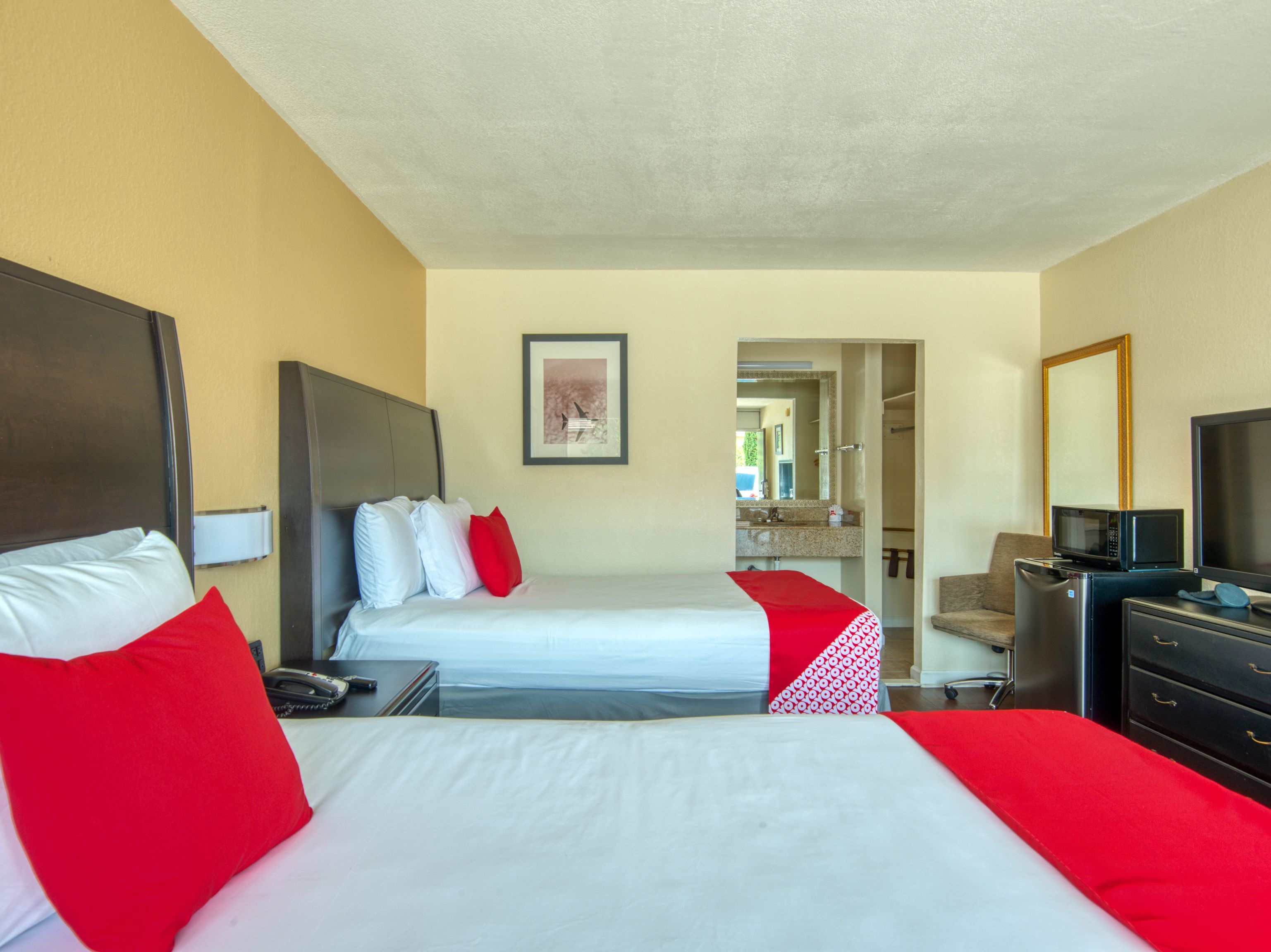 Habitación de hotel con dos camas, un escritorio, una silla, un televisor y un baño visible a través de una puerta abierta.