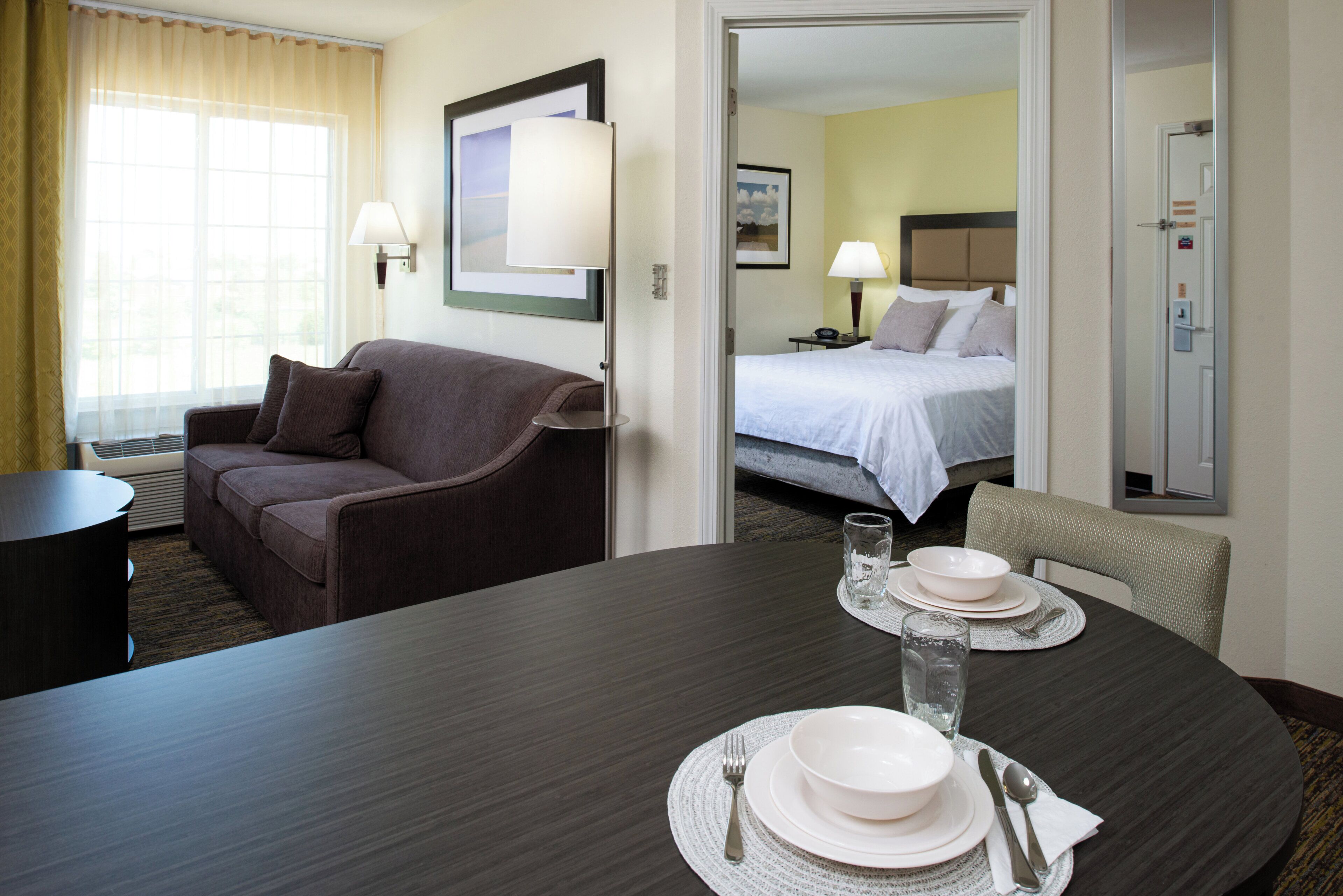 Habitación de hotel con una mesa de comedor lista para dos comensales, un sofá, una cama y un baño visible a través de una puerta abierta.