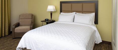 Habitación de hotel con cama, una silla, mesita de noche con lámpara y un reloj de pared.