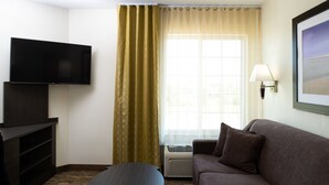Een hotelkamer met een wandgemonteerde flatscreen-tv, een bruine bank, een klein rond tafeltje en een groot raam met gordijnen.