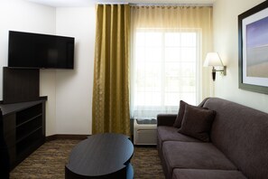 Une chambre d’hôtel dotée d’un téléviseur à écran plat fixé au mur, d’un canapé marron, d’une petite table ronde et d’une grande fenêtre avec des rideaux.
