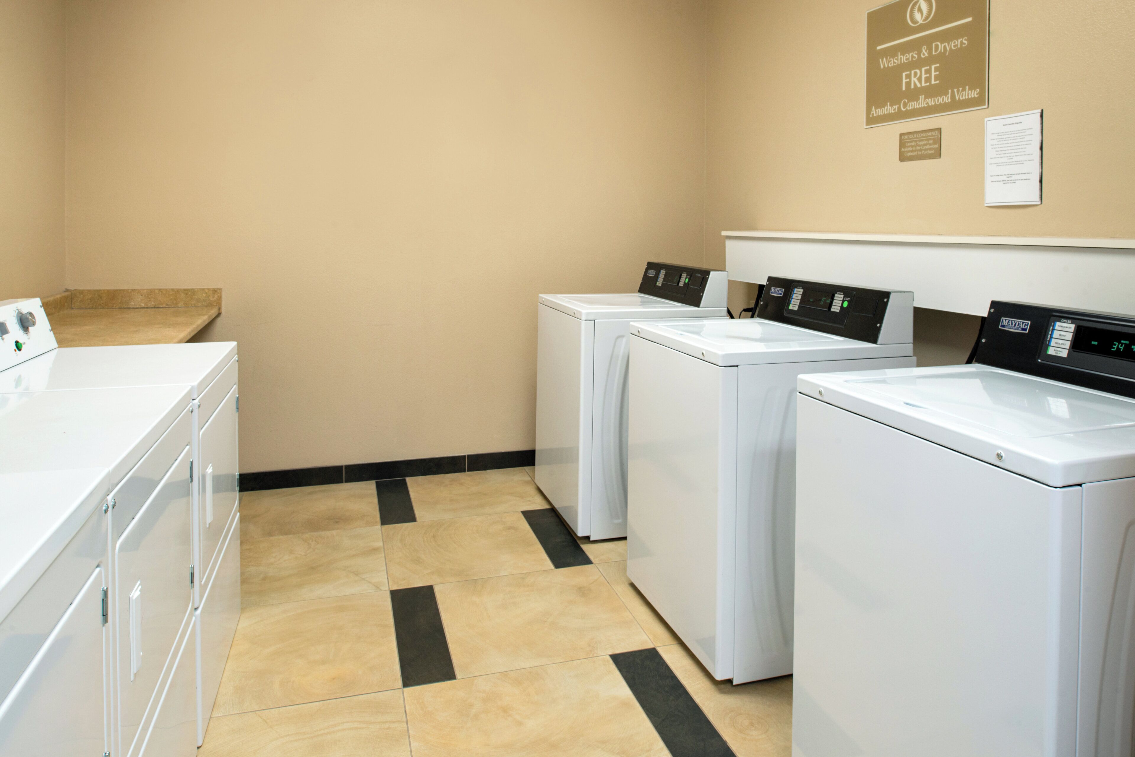 Un cuarto de lavado con tres lavadoras blancas y un panel de control, una pared color beige y piso de baldosas.