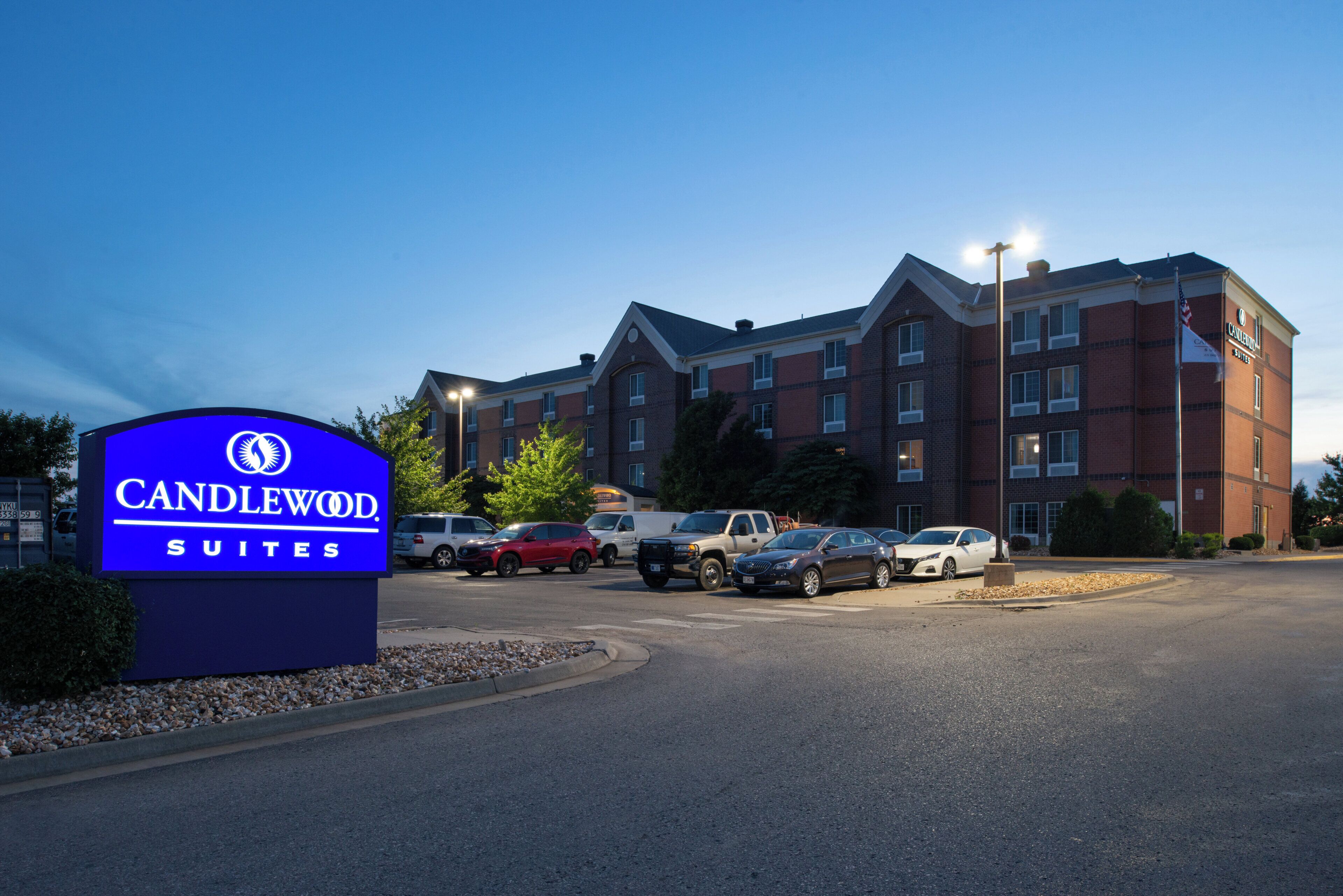 L’entrée d’un hôtel, avec une enseigne indiquant « Candlewood Suites », un parking avec plusieurs voitures et un bâtiment en briques doté de nombreuses fenêtres.