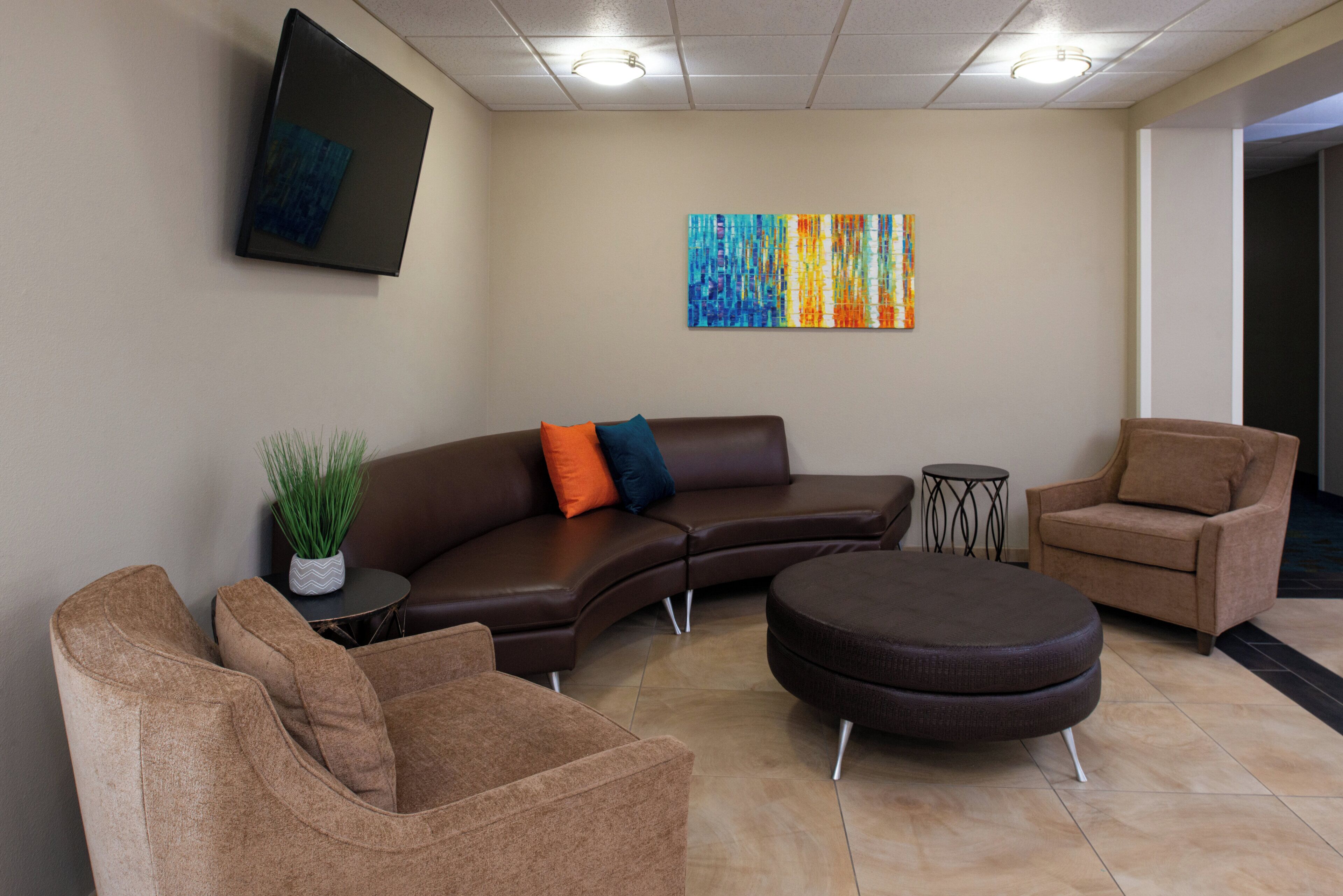 Un espace d’attente moderne comprenant un canapé d’angle, deux fauteuils, un pouf rond, une petite table d’appoint avec une plante, et un tableau abstrait aux couleurs vives accroché au mur.