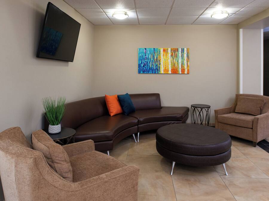 Un espace d’attente moderne comprenant un canapé d’angle, deux fauteuils, un pouf rond, une petite table d’appoint avec une plante, et un tableau abstrait aux couleurs vives accroché au mur.