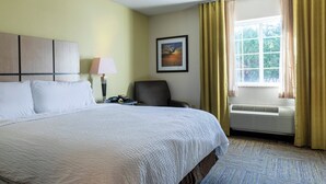 Une chambre d’hôtel avec un lit, une chaise, un bureau avec une lampe, un tableau au mur et une fenêtre avec des rideaux.