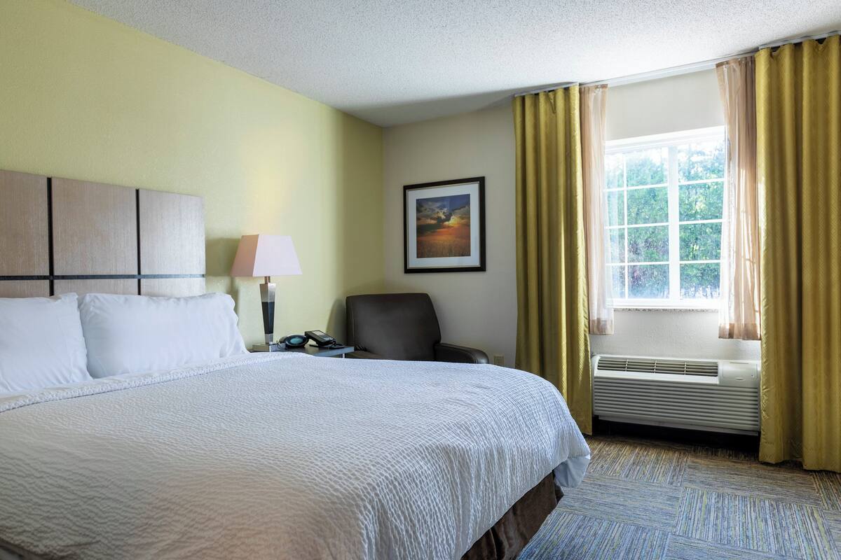 Une chambre d’hôtel avec un lit, une chaise, un bureau avec une lampe, un tableau au mur et une fenêtre avec des rideaux.