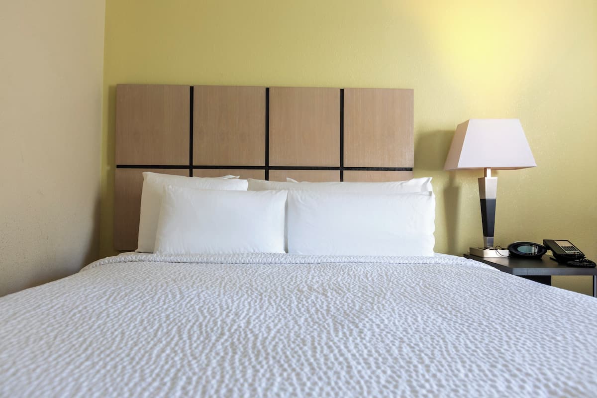 Un lit bien fait, recouvert d’une couvre-lit blanche à texture, avec une tête de lit en bois dotée de panneaux horizontaux, et une table de chevet sur laquelle se trouvent une lampe et un téléphone.