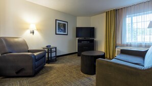 Une chambre d’hôtel équipée d’un canapé, d’un fauteuil, d’une petite table, d’un téléviseur à écran plat et d’une fenêtre avec des rideaux.