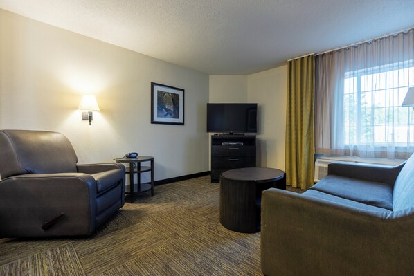 Quarto de hotel com sofá, cadeira, mesinha, televisão de tela plana e janela com cortinas.