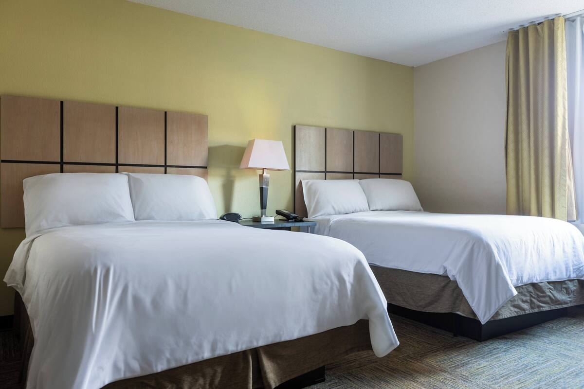 Une chambre d’hôtel avec deux lits doubles, une décoration aux couleurs neutres, une table de chevet avec une lampe et une fenêtre avec des rideaux.