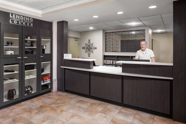A modern hotel reception area with a dark wood reception desk, a large decorative wall piece, and a LENDING LOCKER sign on the left. - Candlewood Suites Omaha Airport by IHG (Omaha)